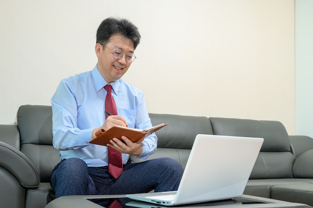 Middle-aged Asian businessman sitting on sofa in living room working at home. Telecommuting concept.