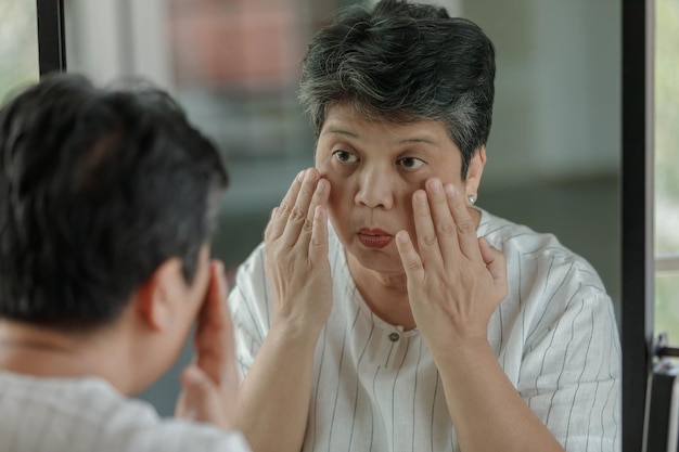 Middle aged Asian female touching face and checking under eye puffiness and wrinkle while looking at mirror at home