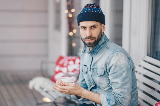 Middle aged bearded male with blue eyes thick beard and mustache wears stylish hat and denim jacket holds cup of hot tea or coffee has thoughtful expression thinks about something important