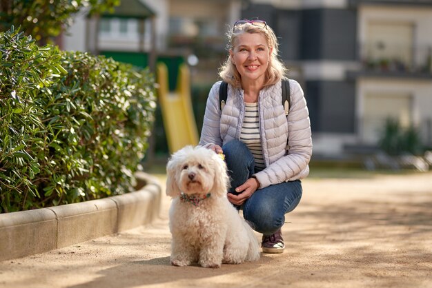 Middle-aged blond woman walking with fluffy white dog in summer city.