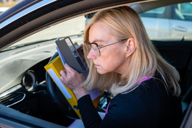 Middle aged blonde business woman in glasses in car working with documents mobile technology concept