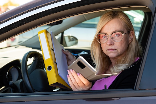 Middle aged blonde business woman in glasses in car working with documents mobile technology concept