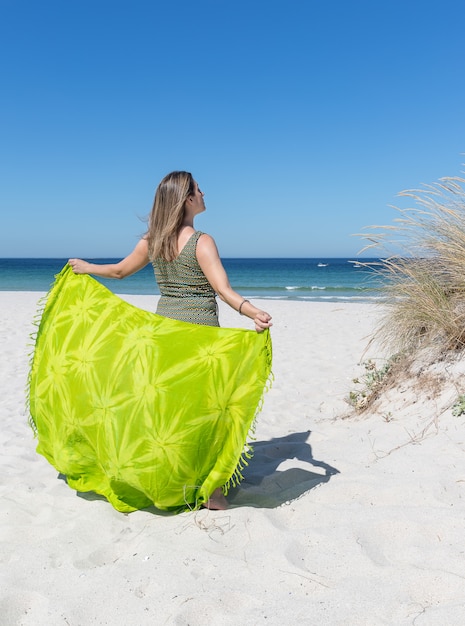 A middle-aged blonde woman on her back holding a green sarong on the beach