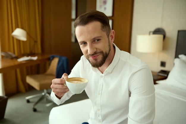 Middle aged businessman, entrepreneur, investment advisor in white shirt sitting on bed in hotel bedroom during business trip, smiles looking at camera, holding a cup with freshly brewed coffee drink