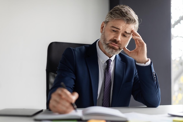 Middle Aged Businessman Sitting At Desk In Office And Writing In Notepad