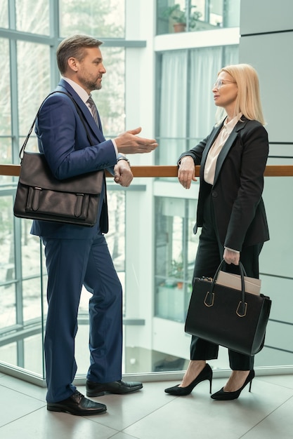 Middle-aged businessman with bag on shoulder gesturing hand while discussing strategy with female colleague in corridor