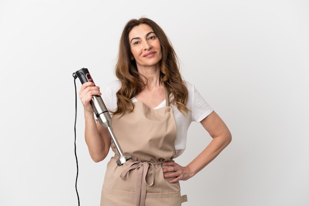 Middle aged caucasian woman using hand blender isolated on white background posing with arms at hip and smiling