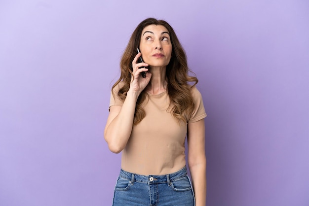 Middle aged caucasian woman using mobile phone isolated on purple background and looking up