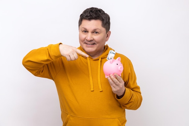 Middle aged man holding pointing at piggy bank with banknote in hands being happy of savings