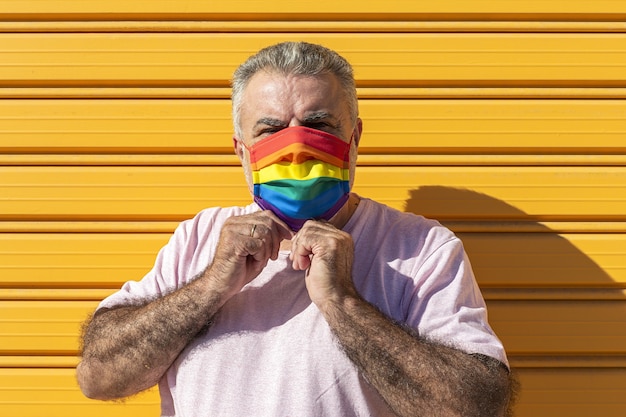 Middle-aged man wearing a hat, sunglasses and a rainbow-colored protective mask. LGTB on yellow background. Covid-19 concept