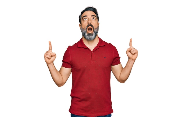 Middle aged man with beard wearing casual red t shirt amazed and surprised looking up and pointing with fingers and raised arms