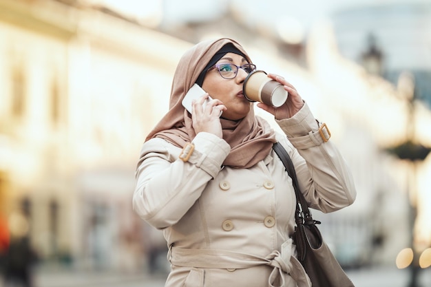 Middle aged Muslim businesswoman wearing hijab with a happy face standing in urban environment, taking on her smartphone and drinking coffee.