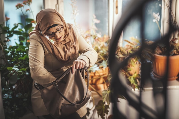 Middle aged Muslim woman wearing hijab with a happy face is standing by the window looking for something in her purse and taking on her smartphone.