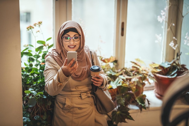 Middle aged Muslim woman wearing hijab with a happy face is standing by the window tipping messages on her smartphone.