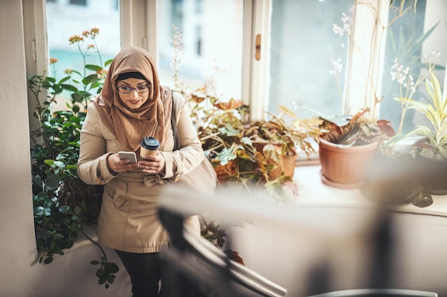 Middle aged Muslim woman wearing hijab with a happy face is standing by the window tipping messages on her smartphone.