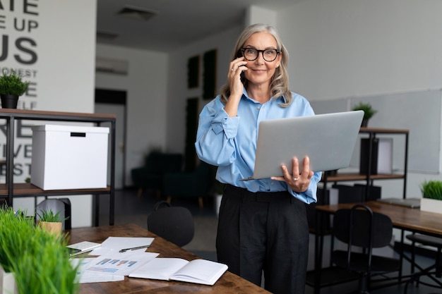 Middle aged older business woman in the office talking on a mobile phone with a laptop in her hands