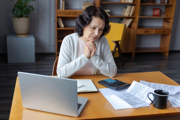 Middle aged senior woman sit with laptop and paper document pensive older mature lady reading paper