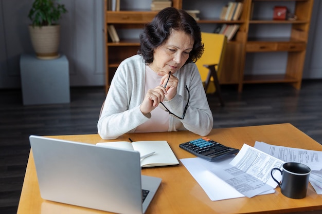 Middle aged senior woman sit with laptop and paper document pensive older mature lady reading paper