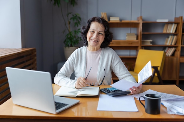 Middle aged senior woman sit with laptop and paper document smiling older mature lady reading paper