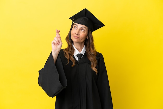 Middle aged university graduate isolated on yellow background with fingers crossing and wishing the best