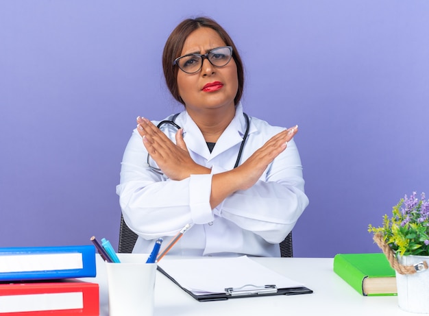 Middle aged woman doctor in white coat with stethoscope wearing glasses looking at front with serious face making stop gesture crossing hands sitting at the table over blue wall