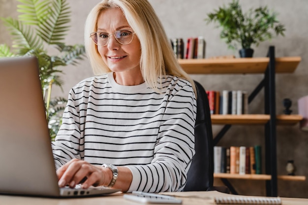 Middle aged woman in eyeglasses sitting at table in office and working on laptop