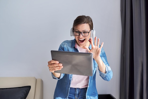 Middle aged woman in headphones with digital tablet at home on armchair