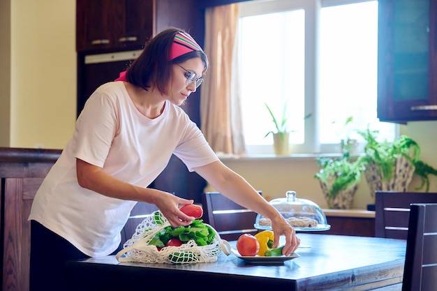 Middle aged woman at home in kitchen with fresh vegetables in mesh textile eco bag