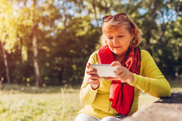 Middle-aged woman relaxing using phone outdoors. Stylish senior lady watching video on smartphone in autumn forest.