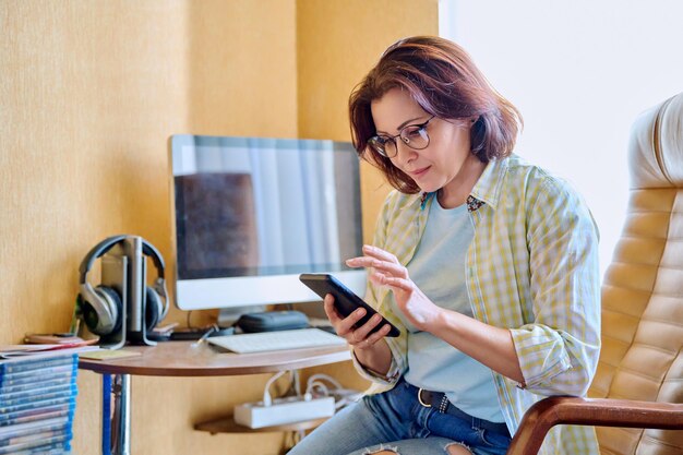 Middle aged woman using smartphone sitting in armchair in home office