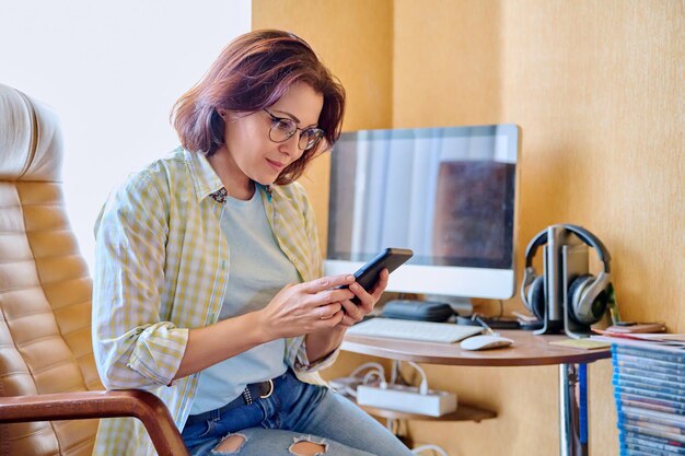 Middle aged woman using smartphone sitting in armchair in home office