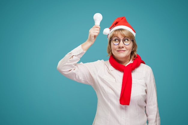 A middle-aged woman in a white shirt, wearing a Christmas hat on a blue background. A woman holds a light bulb, she has an idea.