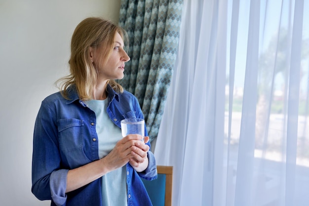 Middle aged woman with glass of water standing at home near window copy space