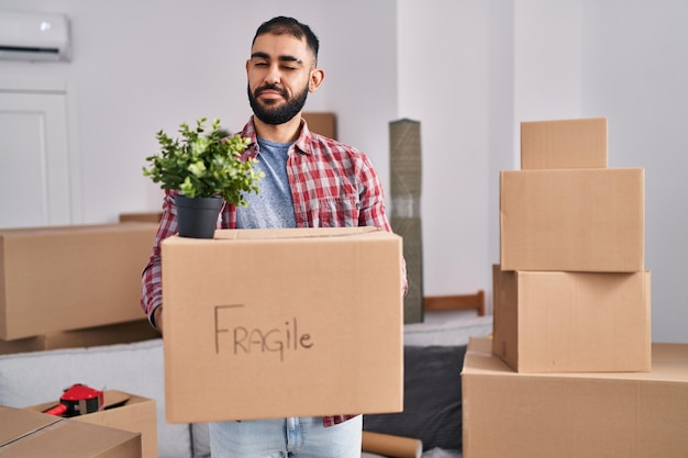 Middle east man with beard moving to a new home holding cardboard box winking looking at the camera with sexy expression, cheerful and happy face.