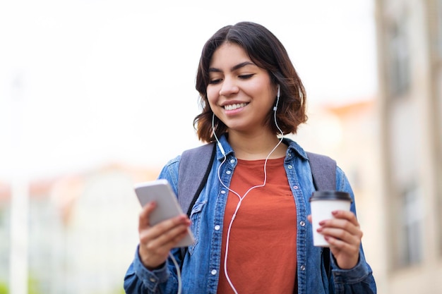 Middle eastern female student listening music on smartphone and drinking coffee outdoors