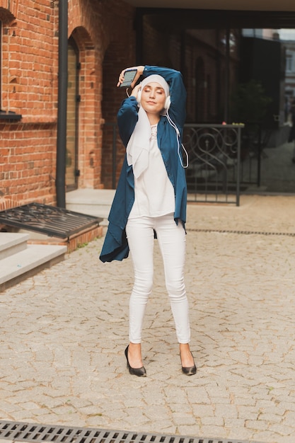 Middle eastern woman in hijab listening music with headphones and dancing outdoors woman