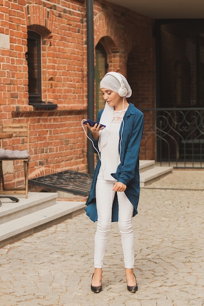 Middle eastern woman in hijab listening music with headphones and dancing outdoors woman