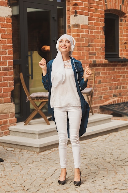 Middle eastern woman in hijab listening music with headphones and dancing outdoors woman