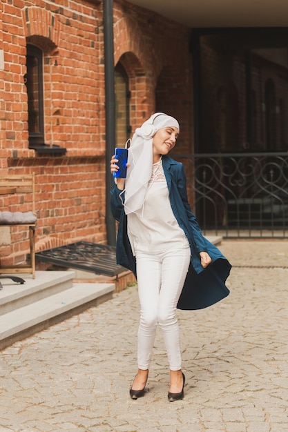 Middle eastern woman in hijab listening music with headphones and dancing outdoors woman