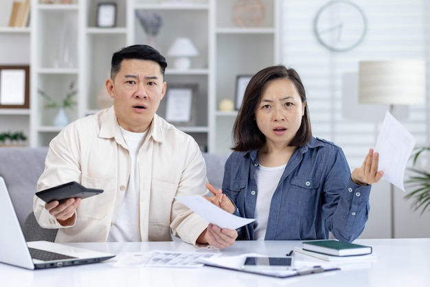 Middleaged asian couple managing documents and budget at home showing confusion and concern laptop