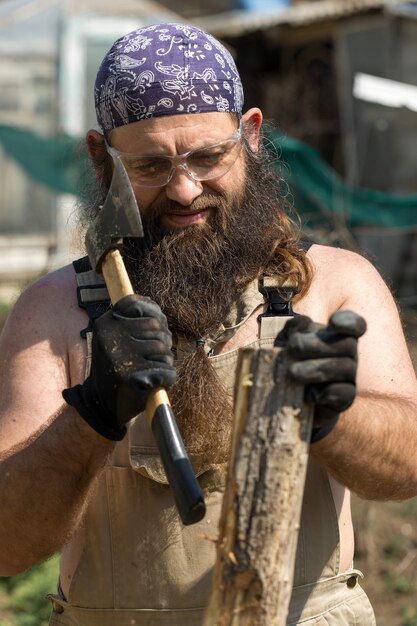 A middleaged bearded man in a bandana cuts logs with an ax Brutal in overalls does the hard work