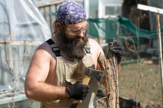 A middleaged bearded man in a bandana cuts logs with an ax Brutal in overalls does the hard work