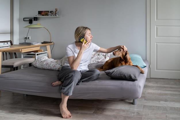 Middleaged blonde woman talks on phone with friend and strokes favourite dog on bed