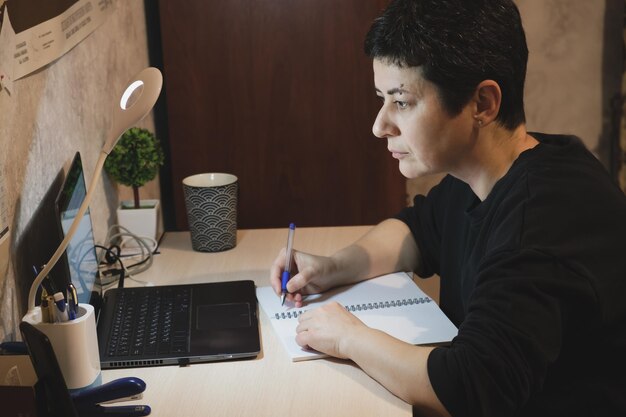 Middleaged brunette woman sits at a desk in front of a laptop screen and makes notes in a notebook