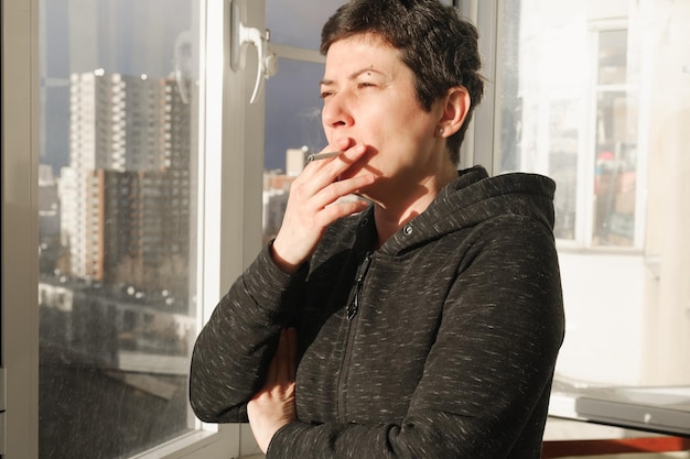 Middleaged brunette woman stands on the glazed balcony of her apartment and smokes