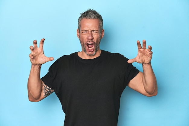 Middleaged caucasian man on blue backdrop screaming with rage