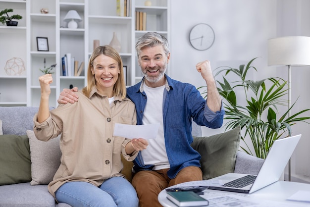 Middleaged couple celebrating while looking at their laptop in a modern living room showing joy and