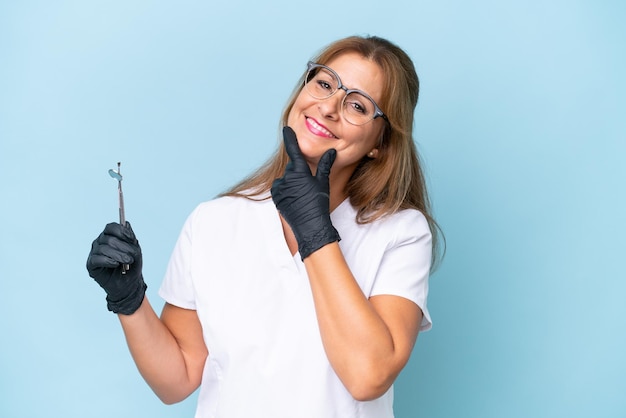 Middleaged Dentist holding tools over isolated blue background happy and smiling