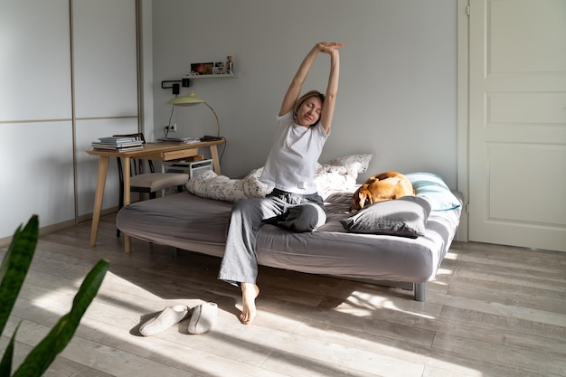 Middleaged female stretches out body sitting on uncovered bed in weekend morning