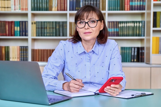 Middleaged female teacher working in library using laptop